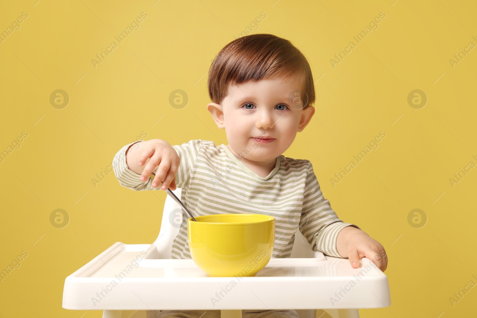 Photo of Cute little kid eating healthy baby food from bowl in high chair on yellow background