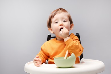 Photo of Cute little kid eating healthy baby food from bowl in high chair on light grey background