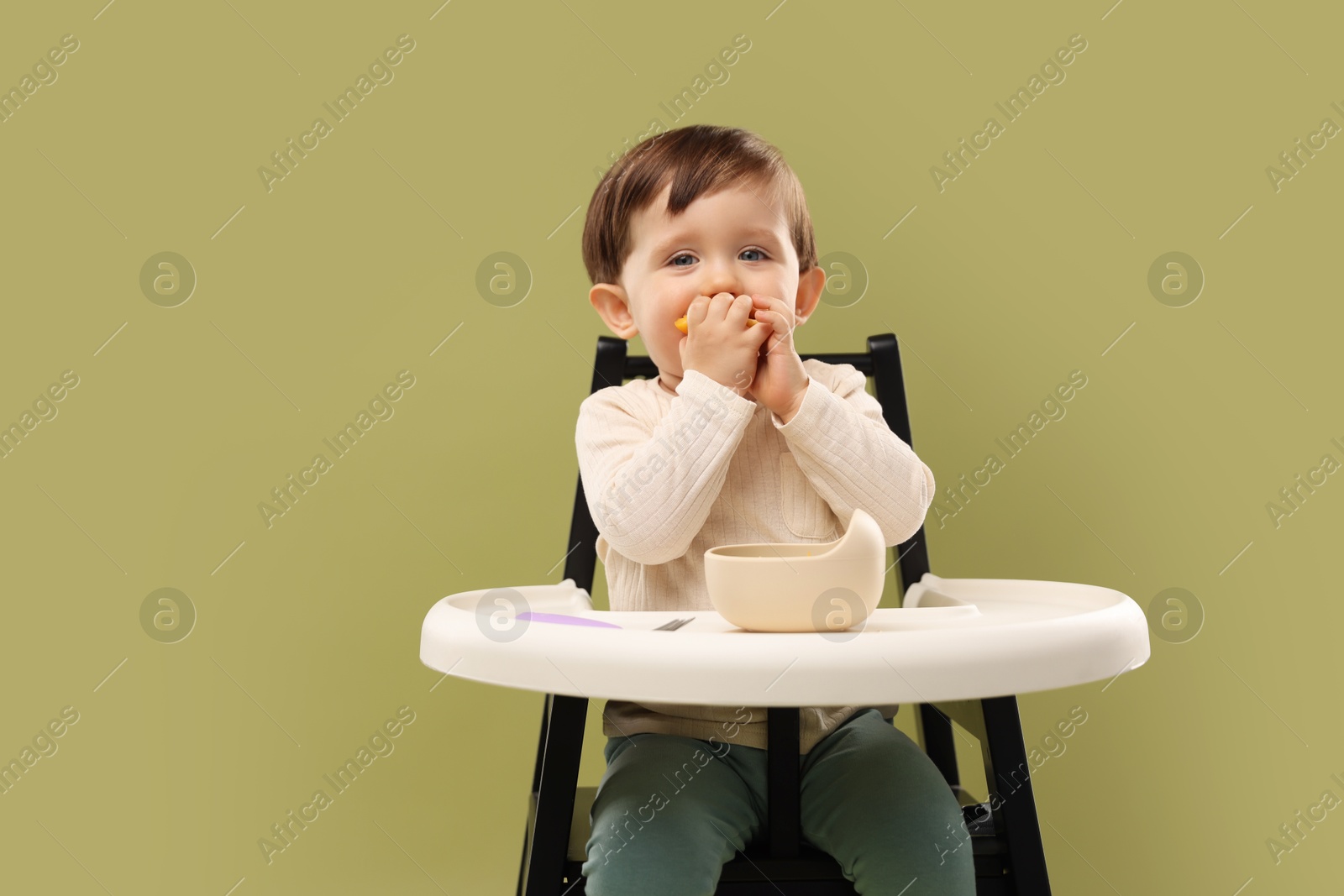 Photo of Cute little baby eating healthy food from bowl in high chair on olive background