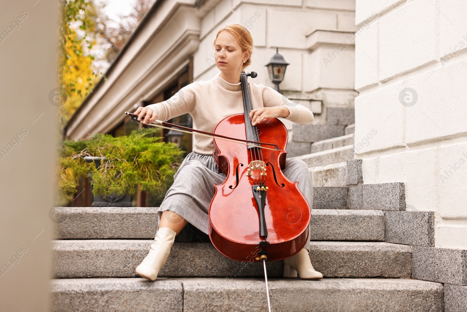 Photo of Beautiful young woman playing cello on stairs outdoors. Classic musical instrument