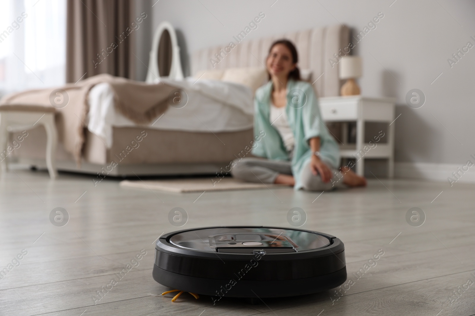 Photo of Young woman sitting in bedroom, focus on robotic vacuum cleaner