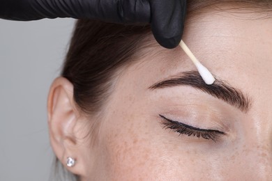 Photo of Brow lamination. Cosmetologist applying cream onto woman's eyebrows against grey background, closeup