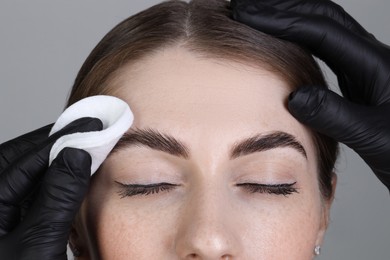 Brow lamination. Cosmetologist wiping woman's eyebrows with cotton pad against grey background, closeup