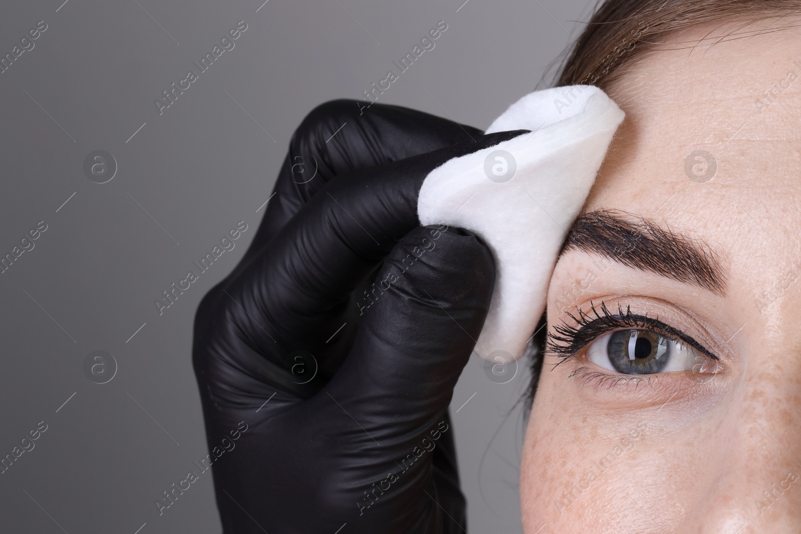 Photo of Brow lamination. Cosmetologist wiping woman's eyebrows with cotton pad against grey background, closeup