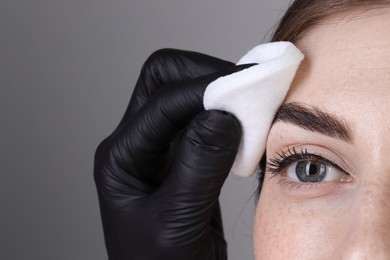 Brow lamination. Cosmetologist wiping woman's eyebrows with cotton pad against grey background, closeup