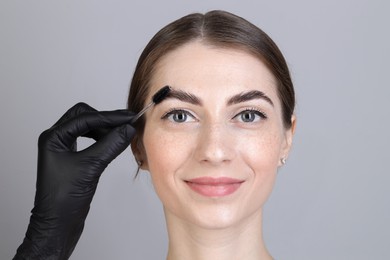 Photo of Brow lamination. Cosmetologist combing woman's eyebrows with brush against grey background, closeup