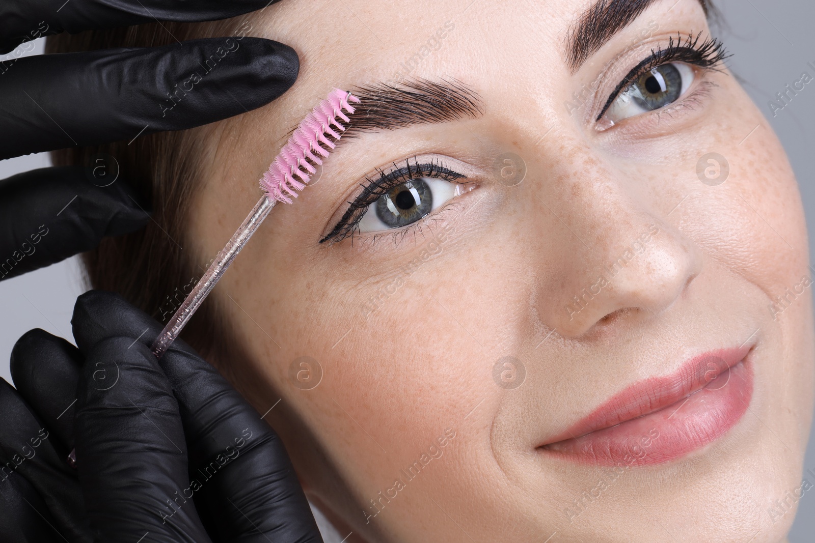 Photo of Brow lamination. Cosmetologist combing woman's eyebrows with brush, closeup