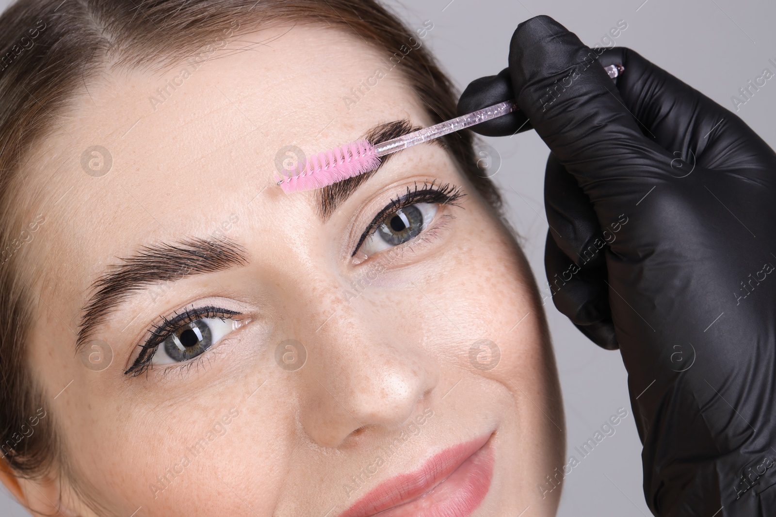 Photo of Brow lamination. Cosmetologist combing woman's eyebrows with brush against grey background, closeup