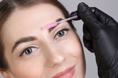 Brow lamination. Cosmetologist combing woman's eyebrows with brush against grey background, closeup