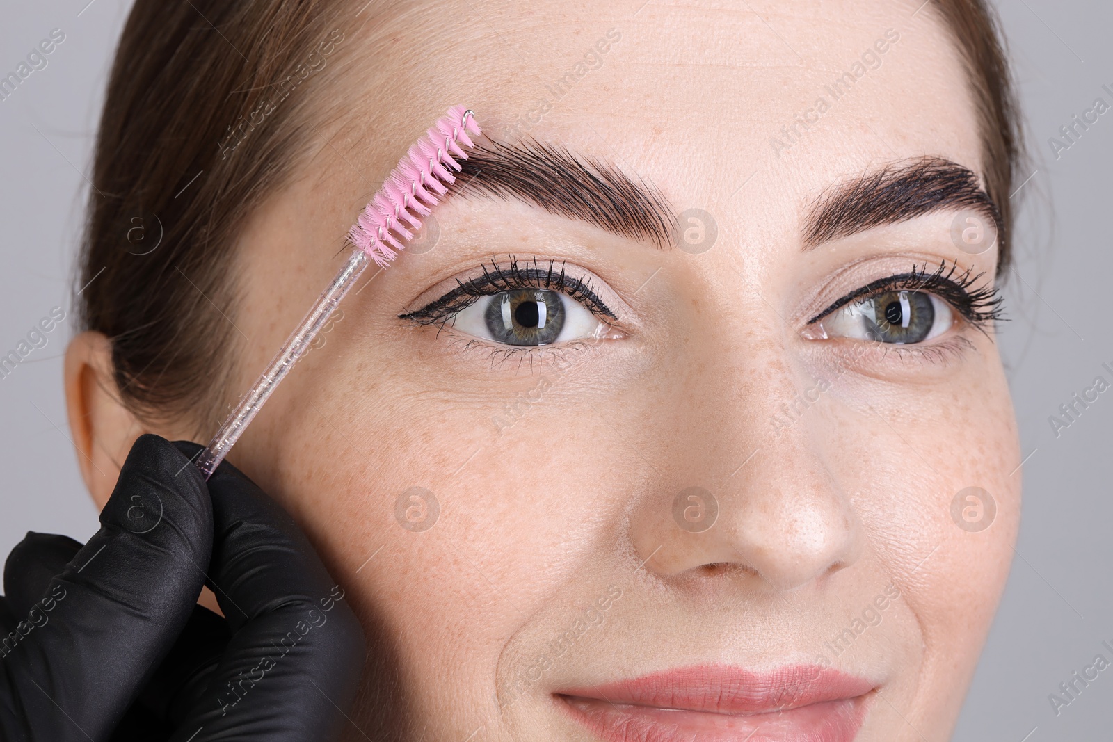 Photo of Brow lamination. Cosmetologist combing woman's eyebrows with brush against grey background, closeup