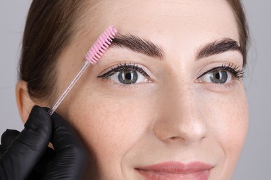 Photo of Brow lamination. Cosmetologist combing woman's eyebrows with brush against grey background, closeup