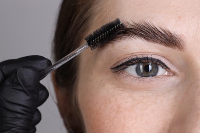 Brow lamination. Cosmetologist combing woman's eyebrows with brush against grey background, closeup