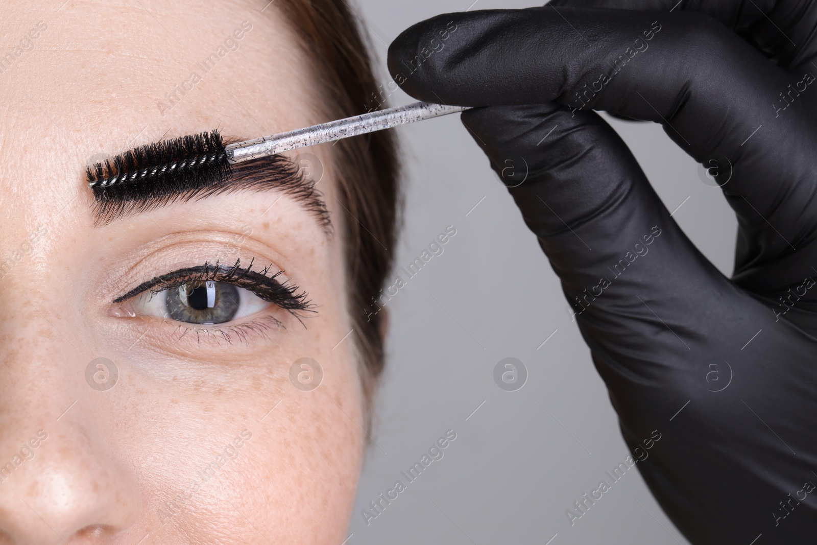 Photo of Brow lamination. Cosmetologist combing woman's eyebrows with brush against grey background, closeup