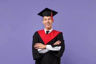 Happy student with crossed arms after graduation on violet background