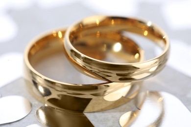 Photo of Beautiful golden wedding rings and heart shaped confetti on light background, closeup