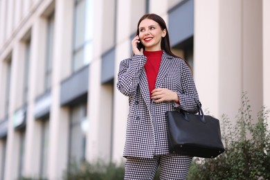 Smiling businesswoman in stylish suit talking on smartphone outdoors, low angle view. Space for text