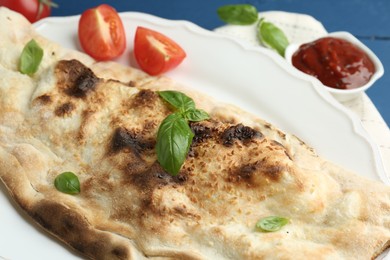 Photo of Tasty calzone with basil, tomatoes and sauce on blue table, closeup