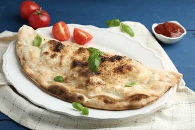 Photo of Tasty calzone with basil, tomatoes and sauce on blue wooden table, closeup