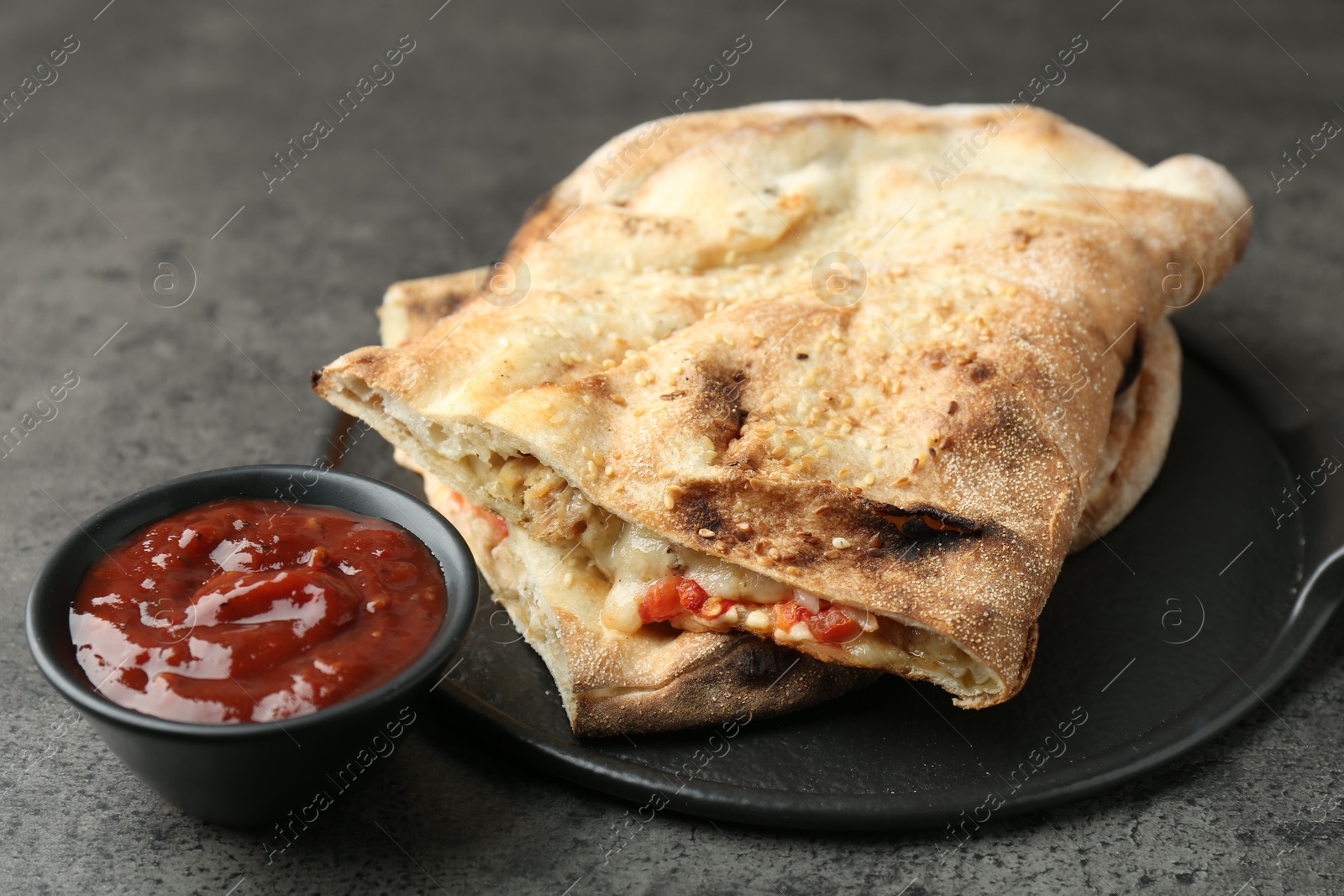 Photo of Halves of tasty calzone with meat, cheese, tomato and sauce on grey textured table, closeup