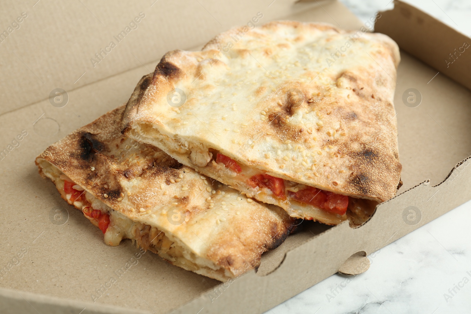 Photo of Halves of tasty calzone with meat, cheese and tomato in cardboard box on white marble table, closeup