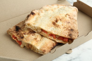 Halves of tasty calzone with meat, cheese and tomato in cardboard box on white marble table, closeup