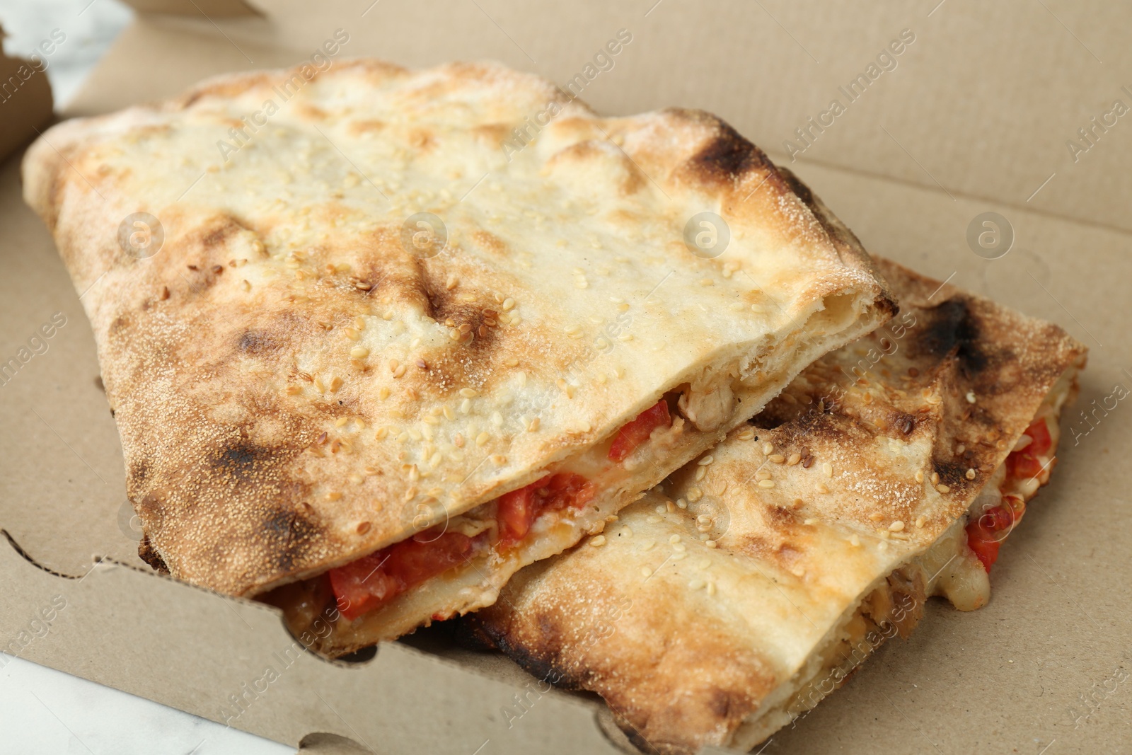 Photo of Halves of tasty calzone with meat, cheese and tomato in cardboard box on white table, closeup