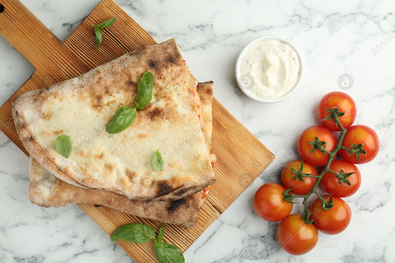 Photo of Halves of tasty calzone, basil, sauce and tomato on white marble table, flat lay