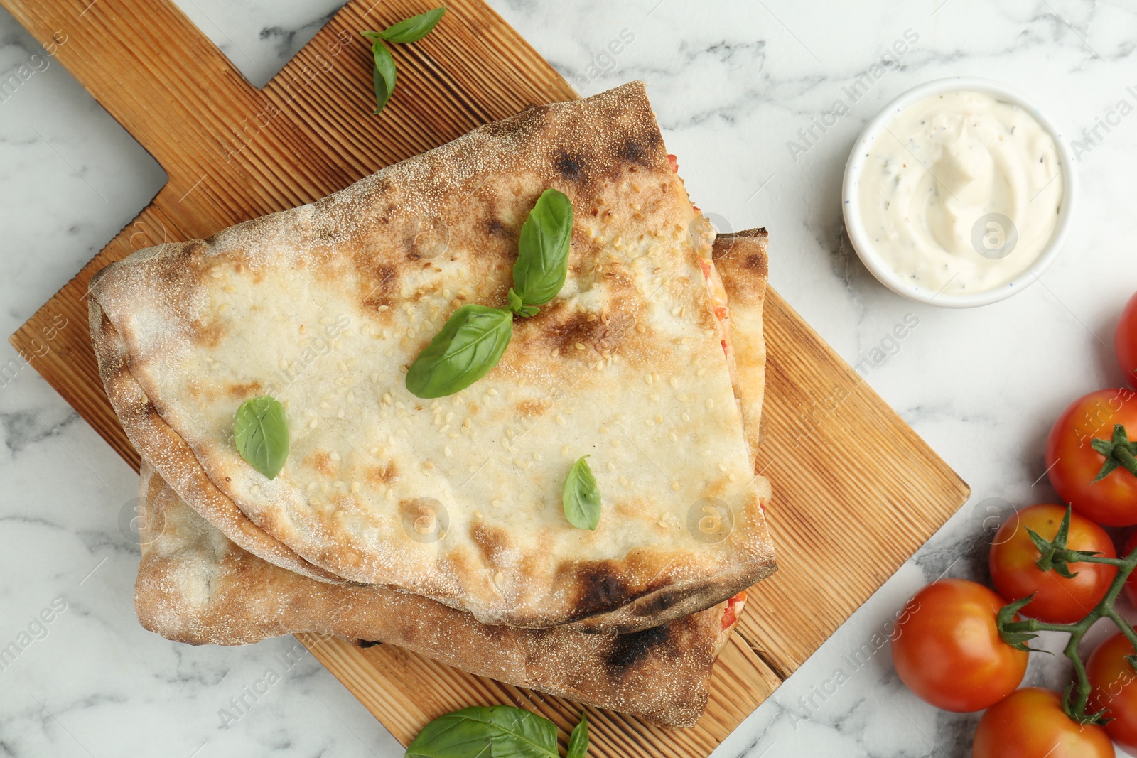 Photo of Halves of tasty calzone, basil, sauce and tomato on white marble table, flat lay