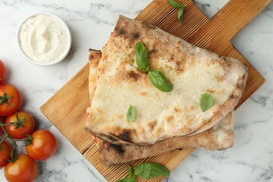 Photo of Halves of tasty calzone, basil, sauce and tomato on white marble table, flat lay