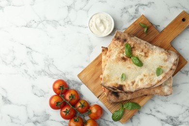 Photo of Halves of tasty calzone, basil, sauce and tomato on white marble table, flat lay. Space for text