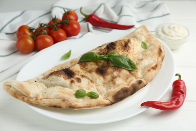 Tasty calzone, basil, chili pepper, sauce and tomatoes on white wooden table, closeup