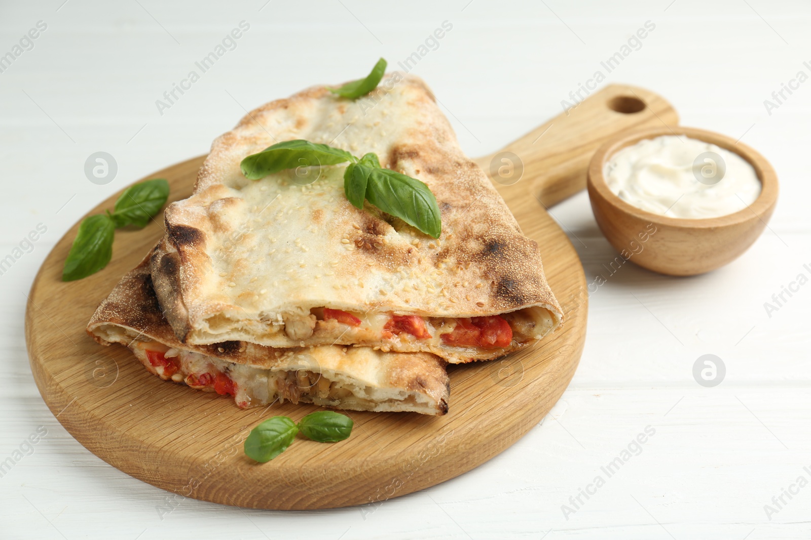 Photo of Halves of tasty calzone with meat, cheese, tomato, basil and sauce on white wooden table, closeup