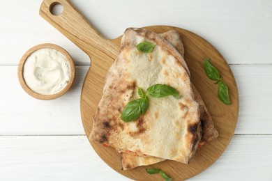 Photo of Halves of tasty calzone, basil and sauce on white wooden table, flat lay