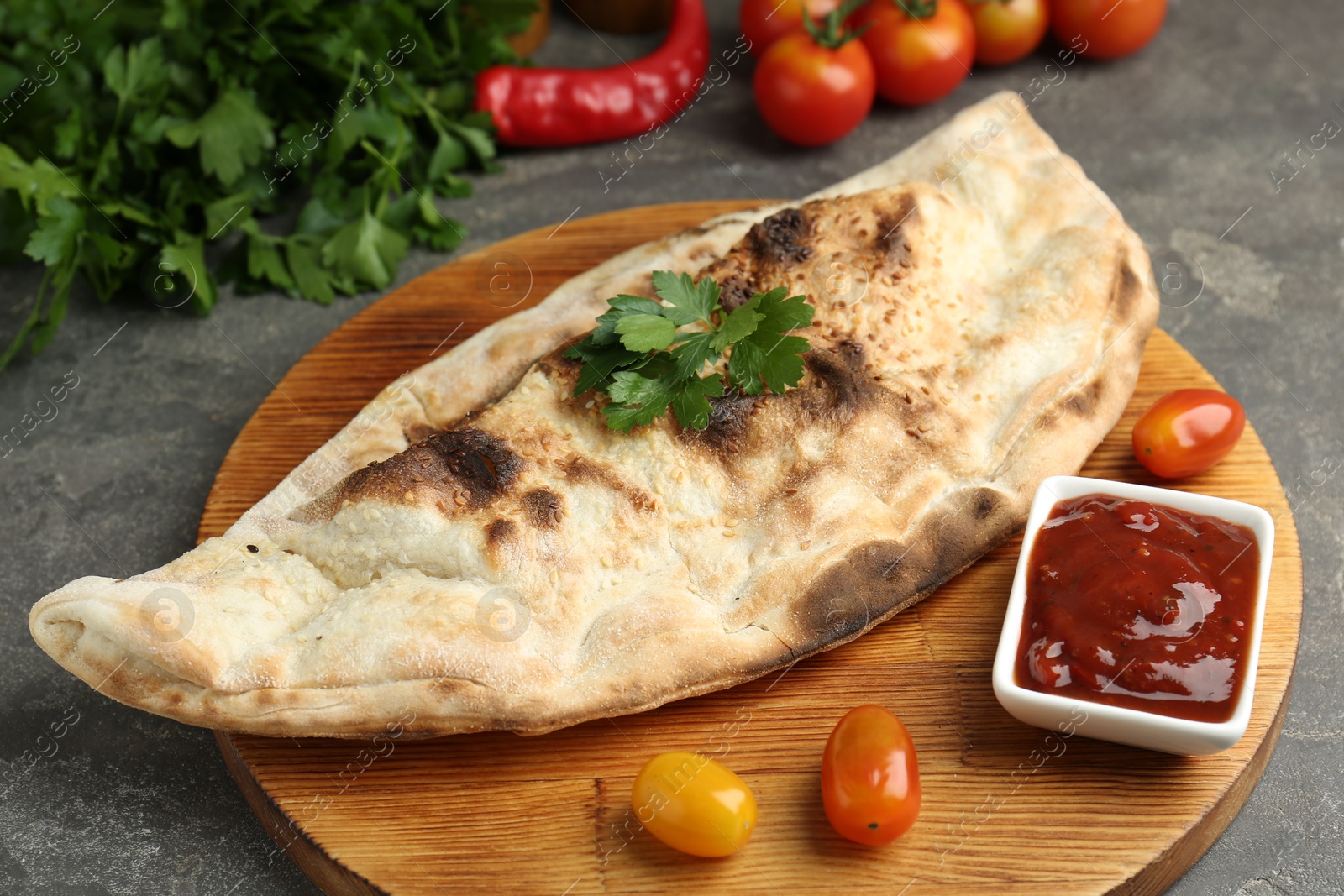 Photo of Board with tasty calzone, tomatoes, sauce and parsley on grey textured table, closeup