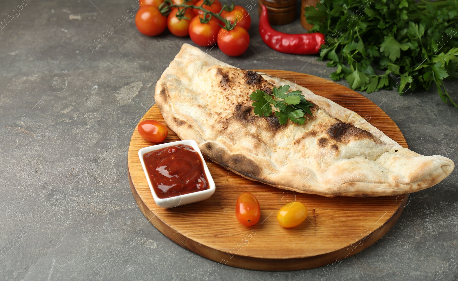 Photo of Board with tasty calzone, tomatoes, sauce and parsley on grey textured table, closeup