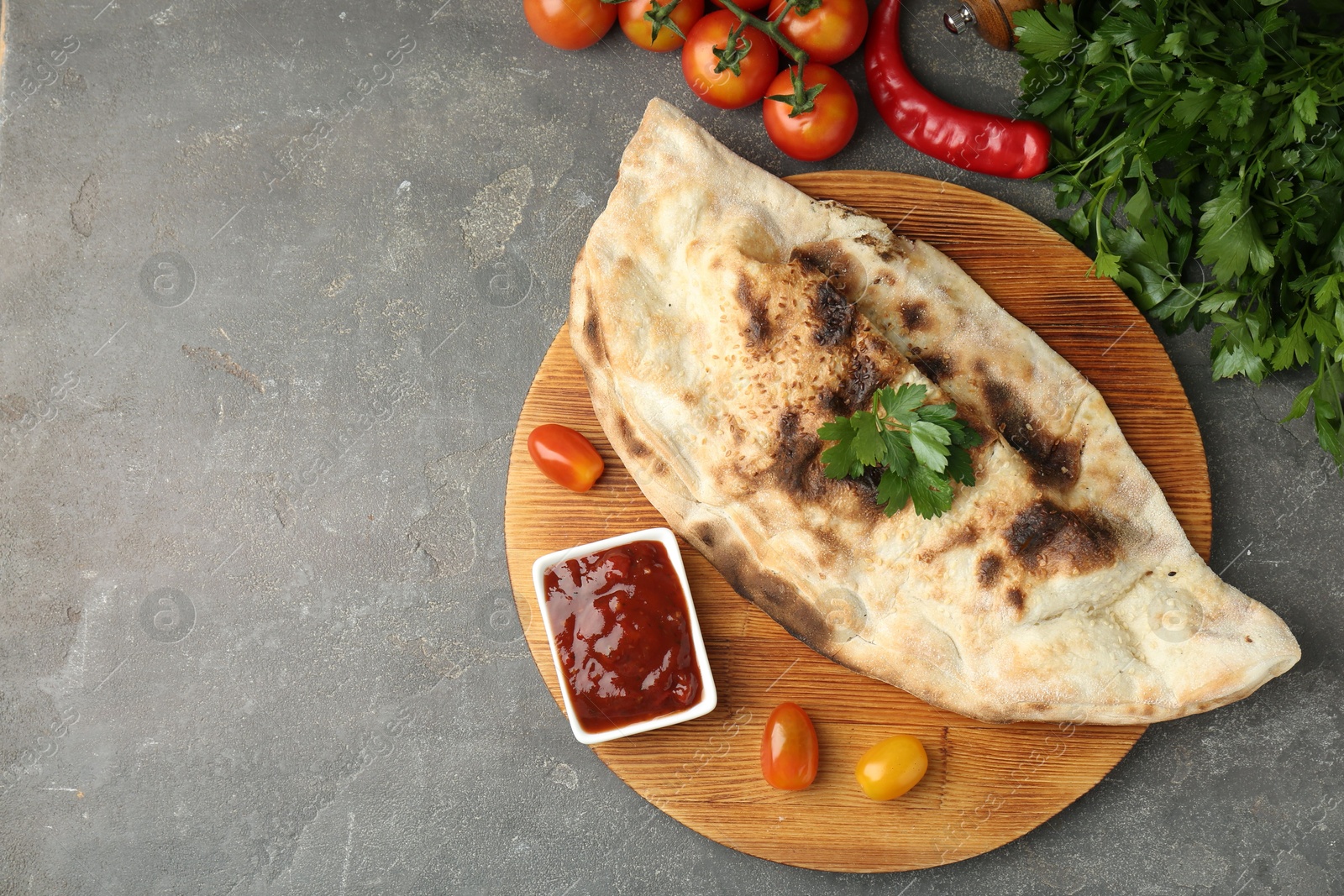 Photo of Board with tasty calzone, tomatoes, sauce and parsley on grey textured table, flat lay. Space for text