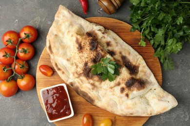 Photo of Board with tasty calzone, tomatoes, sauce and parsley on grey textured table, flat lay