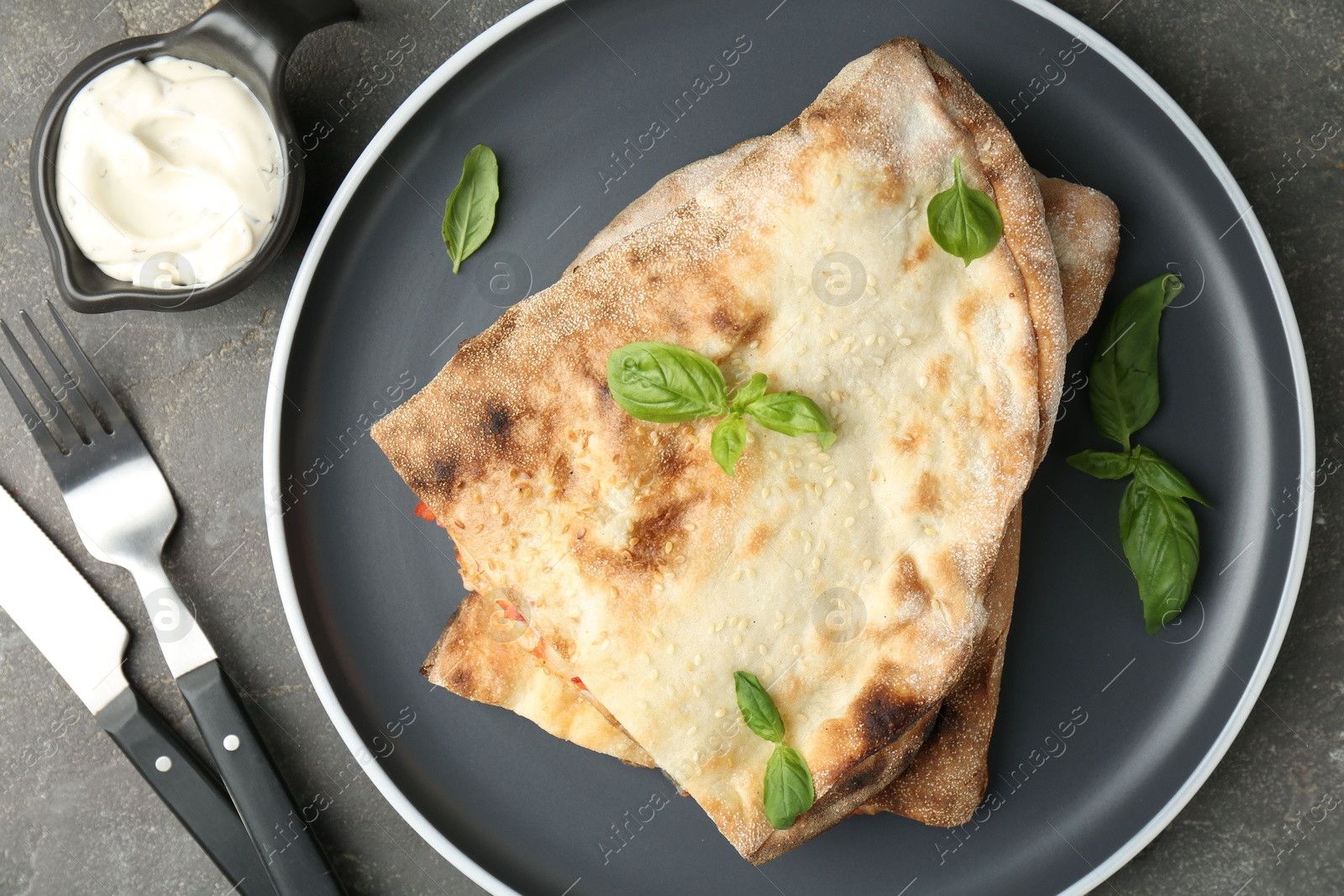 Photo of Halves of tasty calzone with basil served with sauce on grey textured table, flat lay