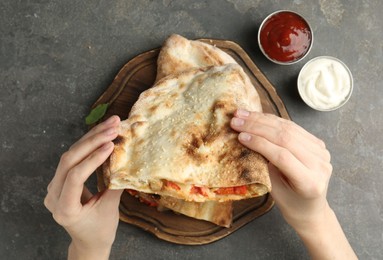 Woman eating tasty calzone with meat, cheese and tomato at grey textured table, top view