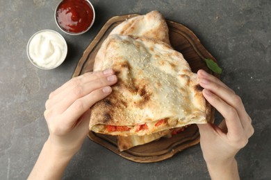 Woman eating tasty calzone with meat, cheese and tomato at grey textured table, top view