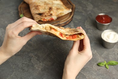 Photo of Woman eating tasty calzone with meat, cheese and tomato at grey textured table, closeup