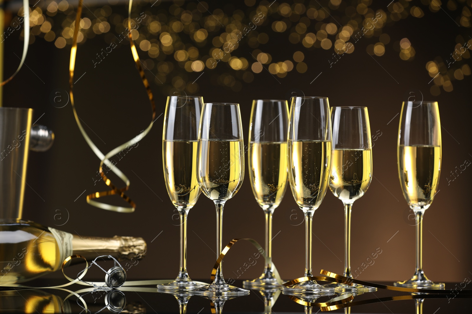 Photo of Champagne in glasses and bottle on mirror surface against brown background with blurred lights