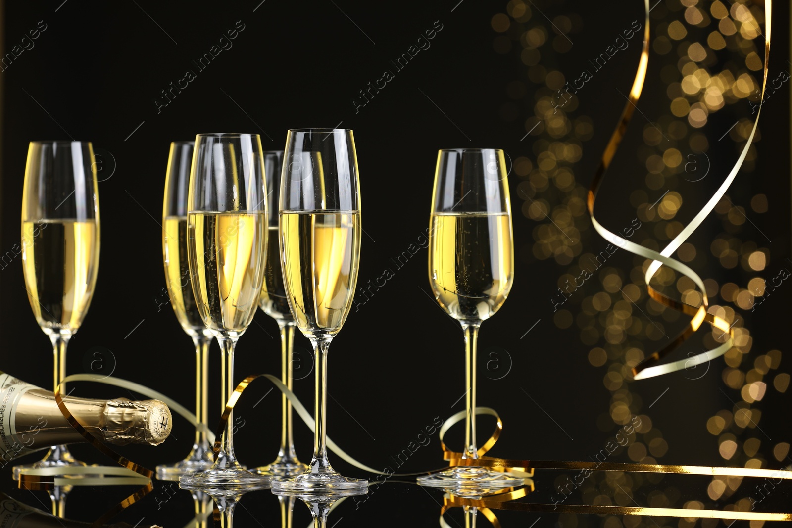 Photo of Champagne in glasses and bottle on mirror surface against black background with blurred lights