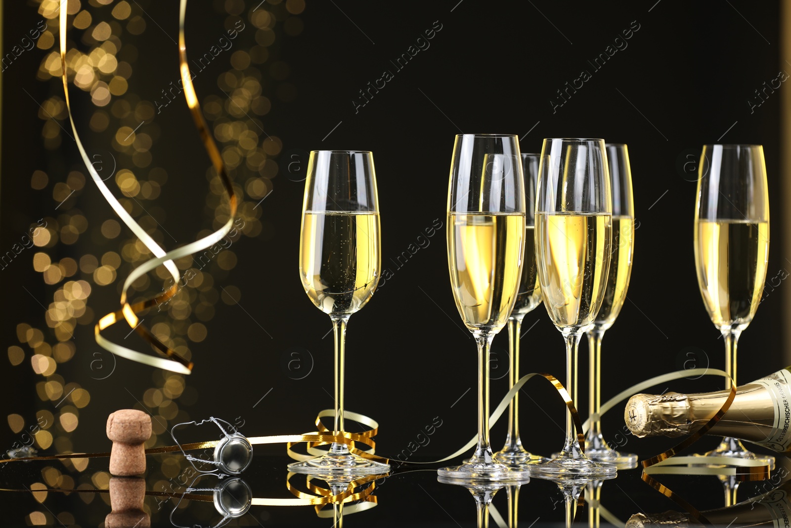 Photo of Champagne in glasses, bottle and cork on mirror surface against black background with blurred lights