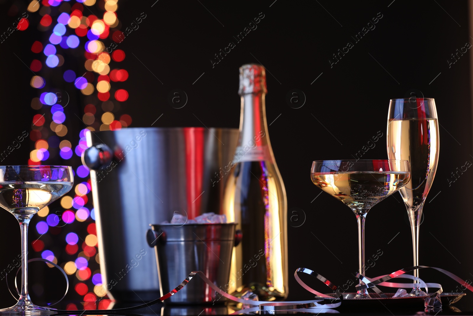 Photo of Champagne in glasses, bottle and ice buckets on mirror surface against black background with blurred lights