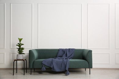 Photo of Stylish sofa with blanket and plant on side table near white wall