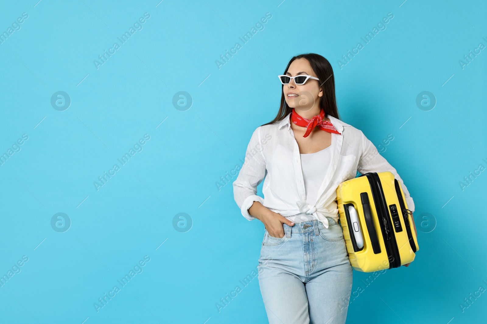 Photo of Happy young woman in sunglasses with suitcase on light blue background, space for text