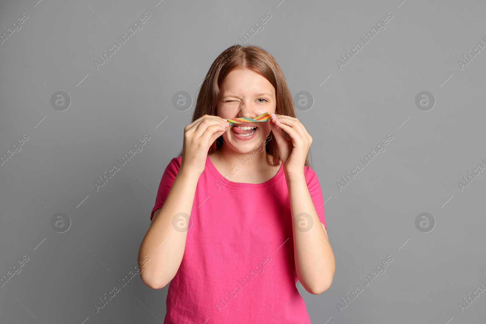 Photo of Happy teenage girl with tasty rainbow sour belt on grey background