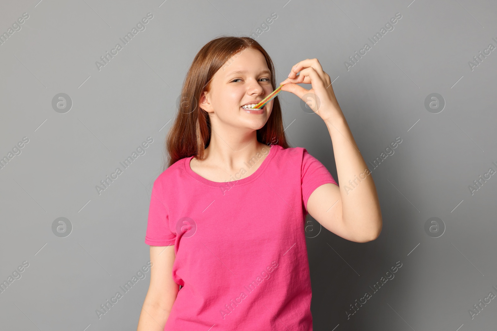 Photo of Teenage girl eating tasty rainbow sour belt on grey background