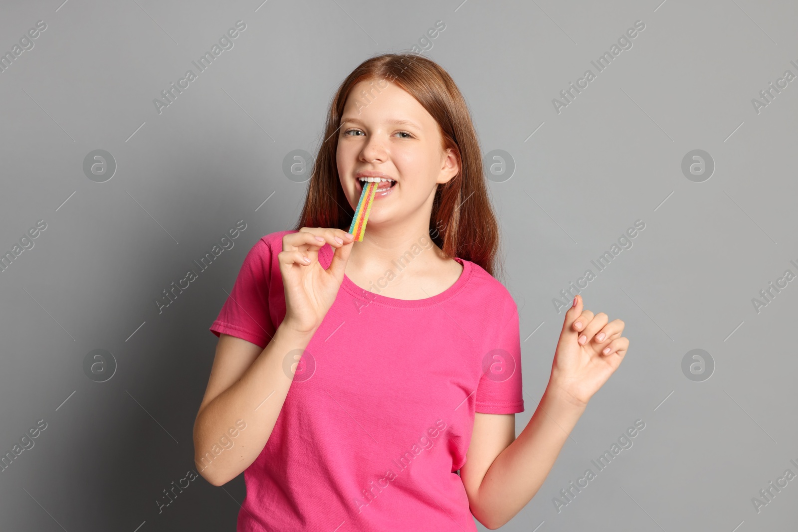 Photo of Teenage girl eating tasty rainbow sour belt on grey background